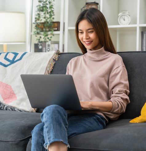 Cheerful young woman sitting on sofa and using laptop computer at home.